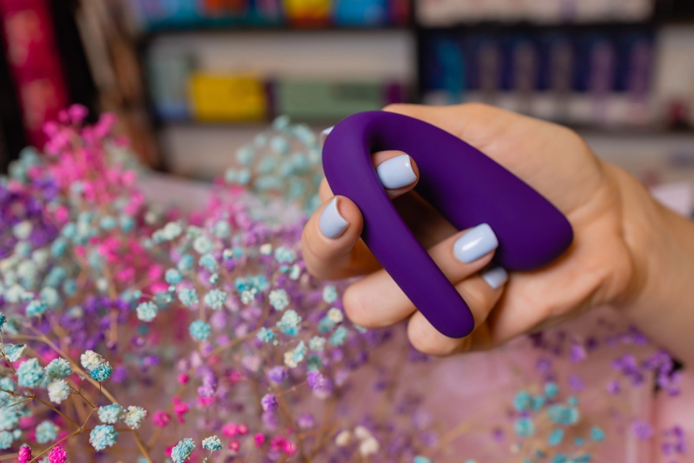 Main pr&eacute;sentant un sextoy de couple dans un love store avec fleurs s&eacute;ch&eacute;es et rayons en fond.